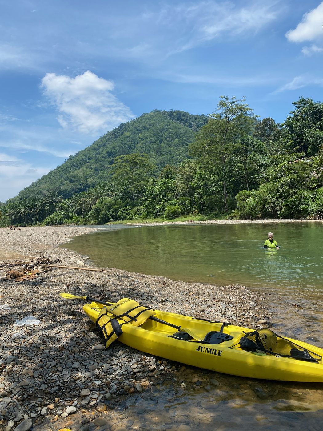 A solo Jungle Kayak boat resting on the riverbank with the lush green mountains of North Sumatra rising behind